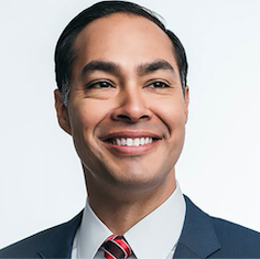Julian Castro wearing a navy suit with a red tie. He is in front of a white background and is looking to the right and smiling.