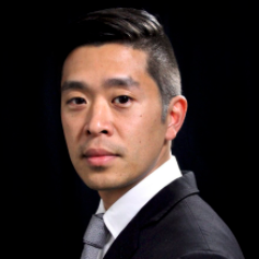 Richard Cho wearing a black suit and grey tie against a black background.