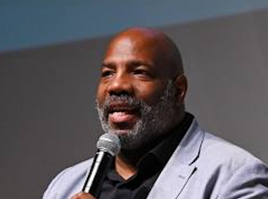 Dr. Jelani Cobb holding a microphone and speaking to an audience. He is wearing a black shirt and a gray suit jacket. 